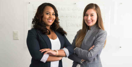 Two women in suits standing beside a wall.