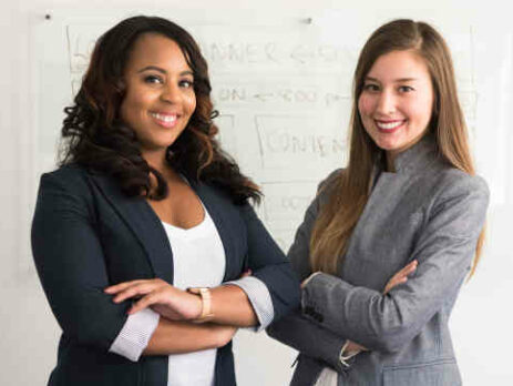 Two women in suits standing beside a wall.