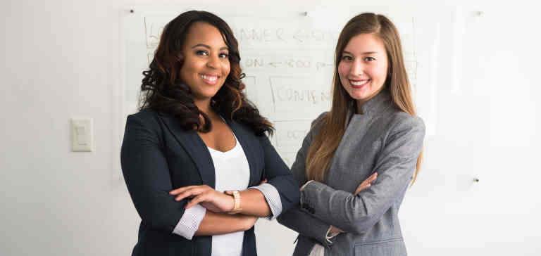 Two women in suits standing beside a wall.