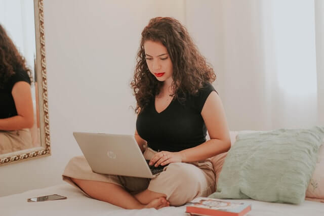 Woman using a laptop while sitting on a bed.