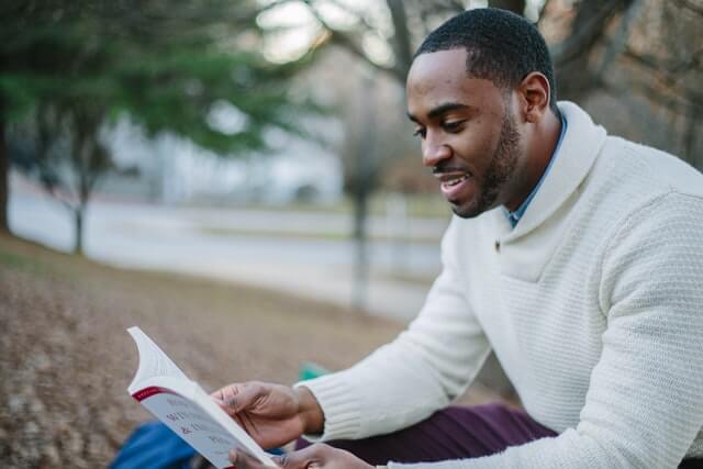 A man wearing a white sweater reads a book in the park.