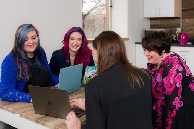 Four women look at a laptop on a brown table.