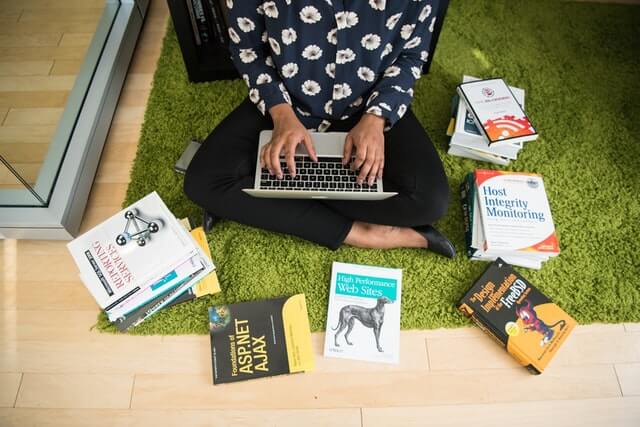 A woman sits among books while working on a laptop.