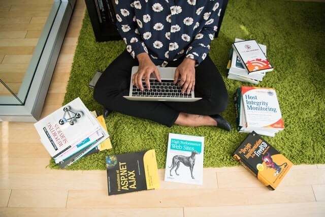 A woman sits among books while working on a laptop.