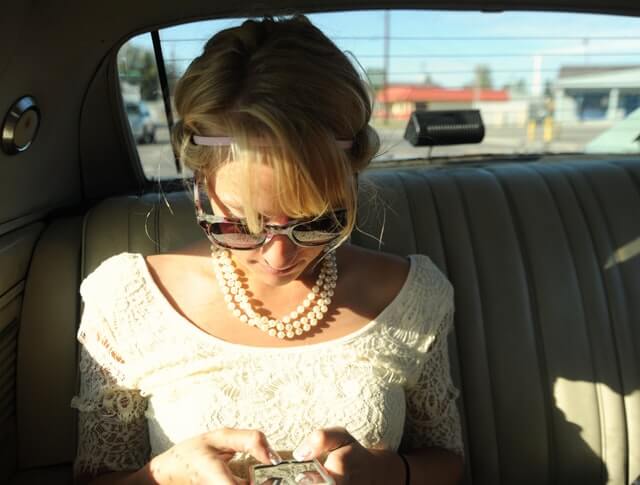 A woman in a white shirt types on her phone in the back seat of a car.