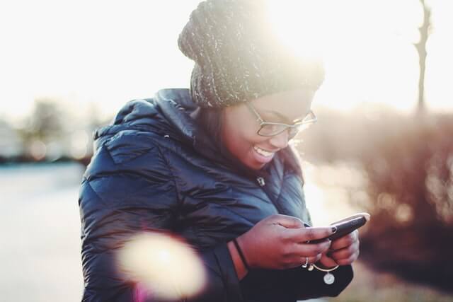 A smiling young woman in a black coat and green hat uses a smartphone.