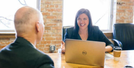 A woman with a laptop talks with a man at a brown table.