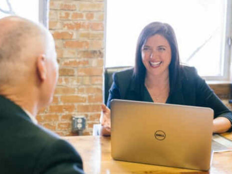 A woman with a laptop talks with a man at a brown table.