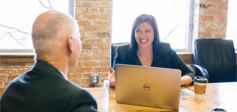 A woman with a laptop talks with a man at a brown table.