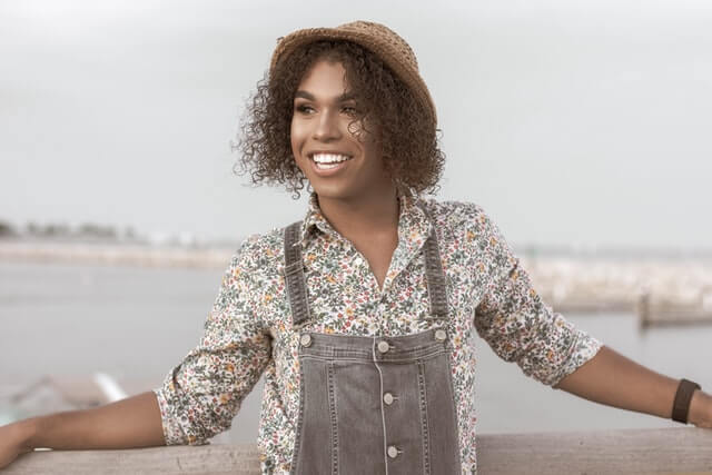 A woman wearing gray overalls and a hat stands near a handrail.