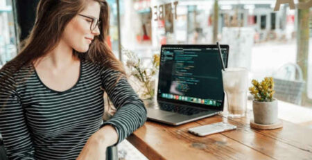 A woman in glasses sits in a coffee shop with a cell phone and laptop.