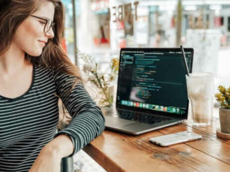 A woman in glasses sits in a coffee shop with a cell phone and laptop.