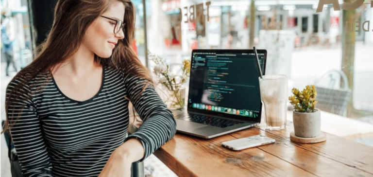 A woman in glasses sits in a coffee shop with a cell phone and laptop.