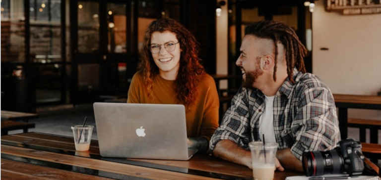 A man and a woman sit at a table with a laptop and a camera.