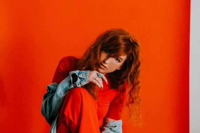 A redheaded woman in red kneels against a red background.