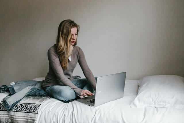 A blonde woman works on a laptop on a bed.