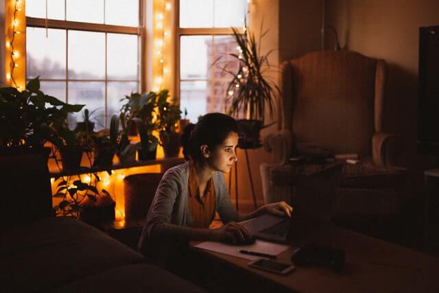 A woman wearing a grey sweater works on a laptop near a window.