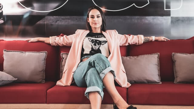 A woman in a pink jacket stretches her arms out on a red couch.