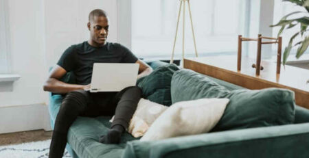 A man wearing a black shirt sits on a couch and works on a laptop.