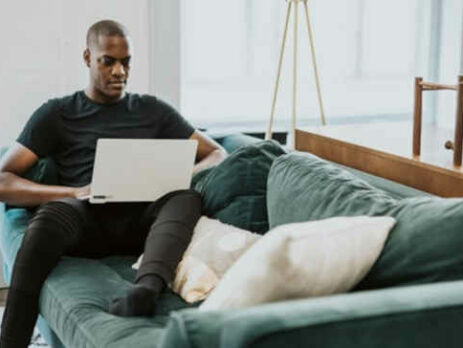 A man wearing a black shirt sits on a couch and works on a laptop.