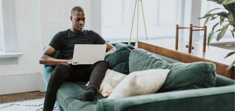 A man wearing a black shirt sits on a couch and works on a laptop.