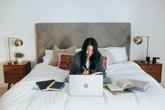 A woman sits on a bed with books and a laptop.