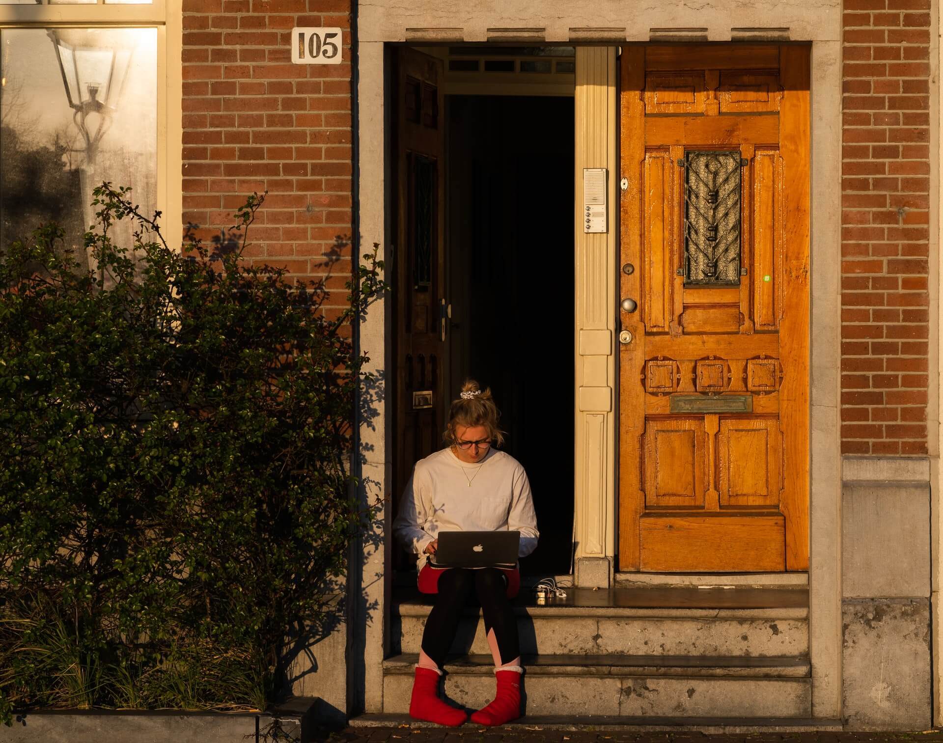 A woman in red socks and a grey sweater works on a laptop outside.