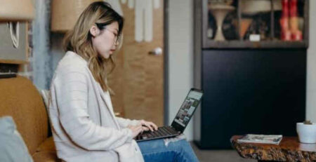 A woman wearing a white sweater works on a laptop at home.