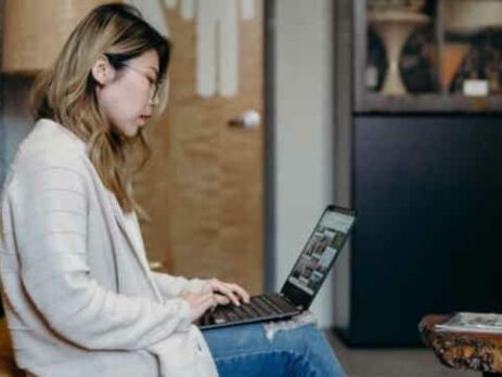 A woman wearing a white sweater works on a laptop at home.