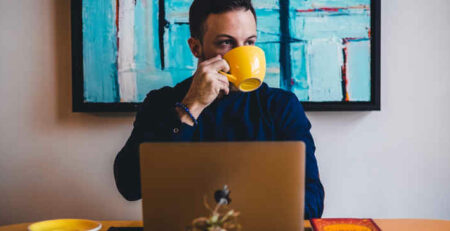 A man drinks coffee from a yellow mug while working on a laptop.