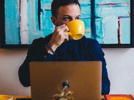 A man drinks coffee from a yellow mug while working on a laptop.