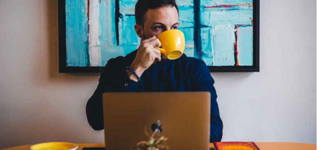 A man drinks coffee from a yellow mug while working on a laptop.