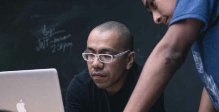 Two men work on a Macbook laptop.