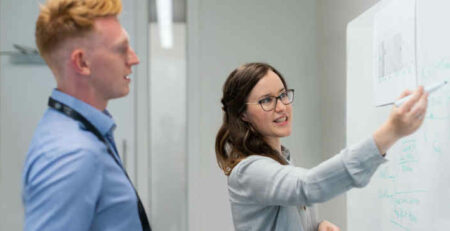 A man and woman talk in front of a whiteboard.