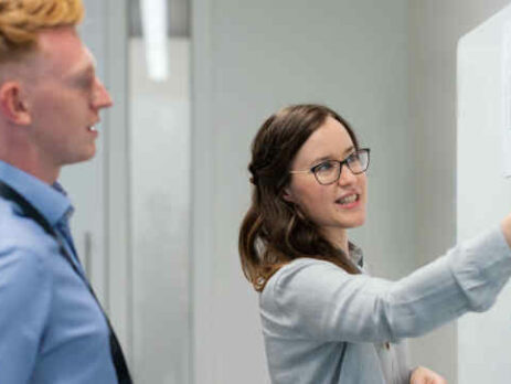 A man and woman talk in front of a whiteboard.