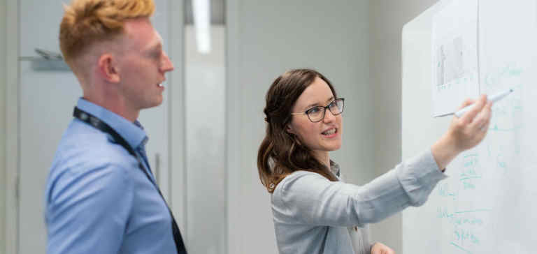 A man and woman talk in front of a whiteboard.