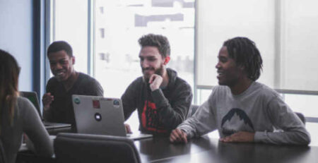 Three men with laptops sit across from a woman at a table.