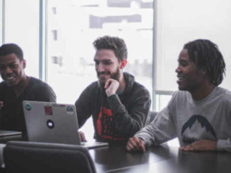 Three men with laptops sit across from a woman at a table.