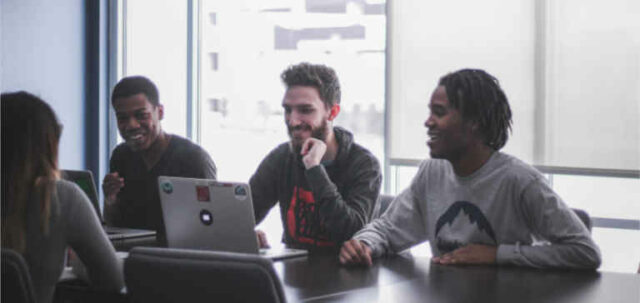 Three men with laptops sit across from a woman at a table.