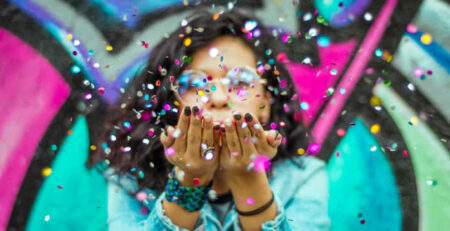 Woman in blue top blows confetti at the camera.