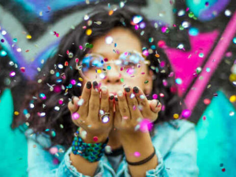 Woman in blue top blows confetti at the camera.