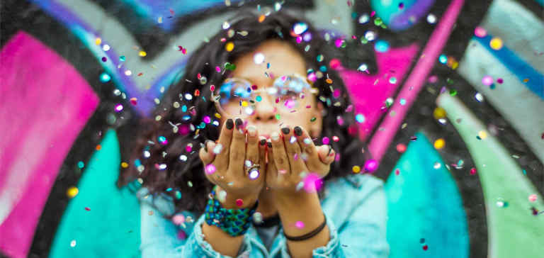 Woman in blue top blows confetti at the camera.