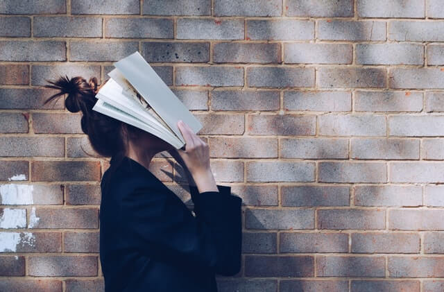 A girl holds a book over her face near a brick wall.