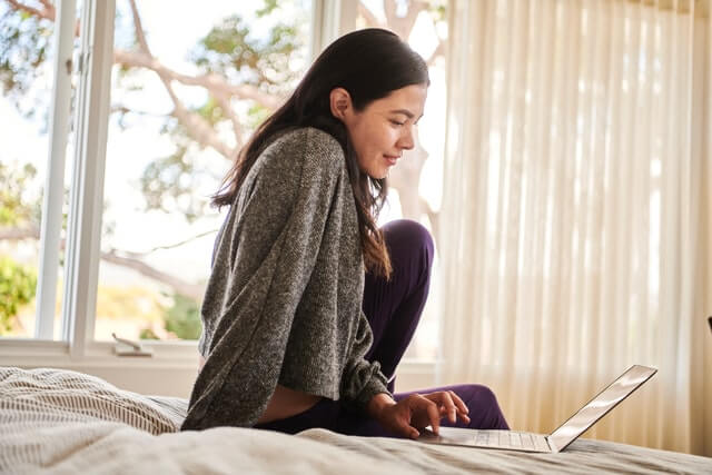 A woman wearing a grey sweater works on a laptop from her bed.