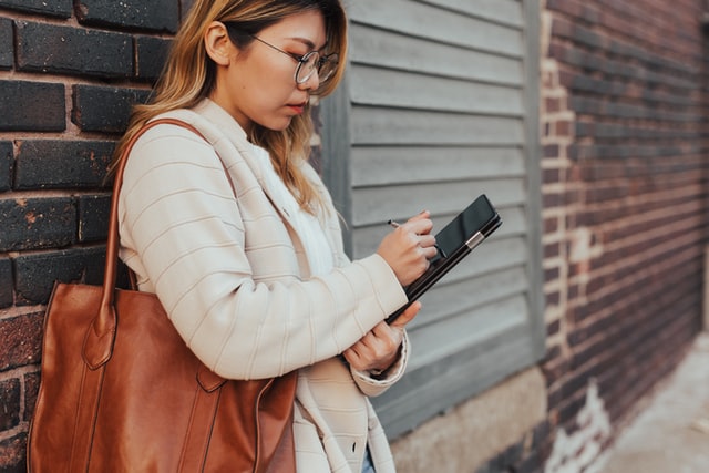 A woman stands against a wall and works on a tablet.