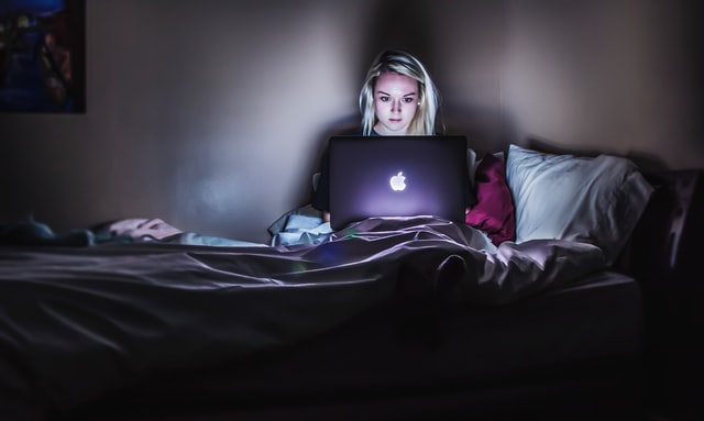 A girl sits on her bed and works on a laptop computer in the dark.
