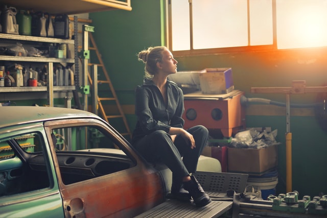 A woman sits on an orange car in a garage.