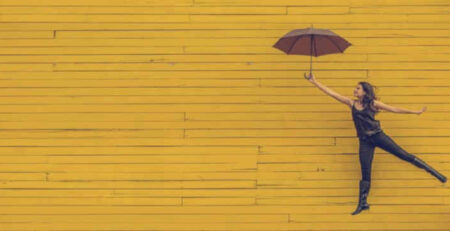 A woman holds a black umbrella near a yellow wall.