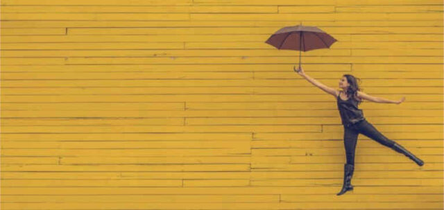 A woman holds a black umbrella near a yellow wall.