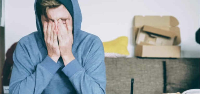 A man in a blue sweater covers his face with his hands.